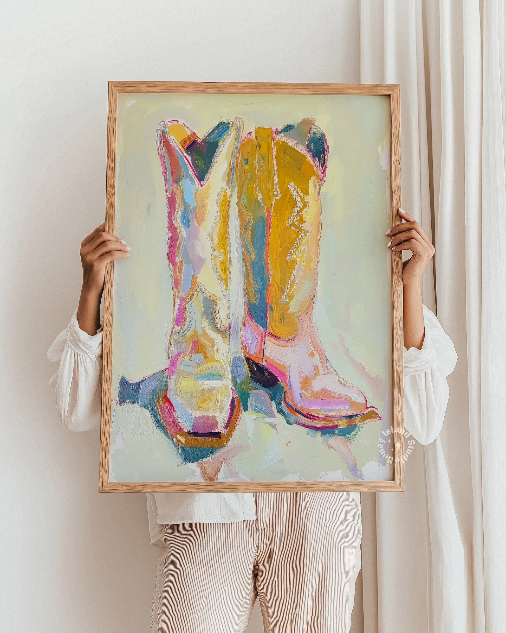 Person holding a framed painting of colorful cowgirl boots against a white wall.