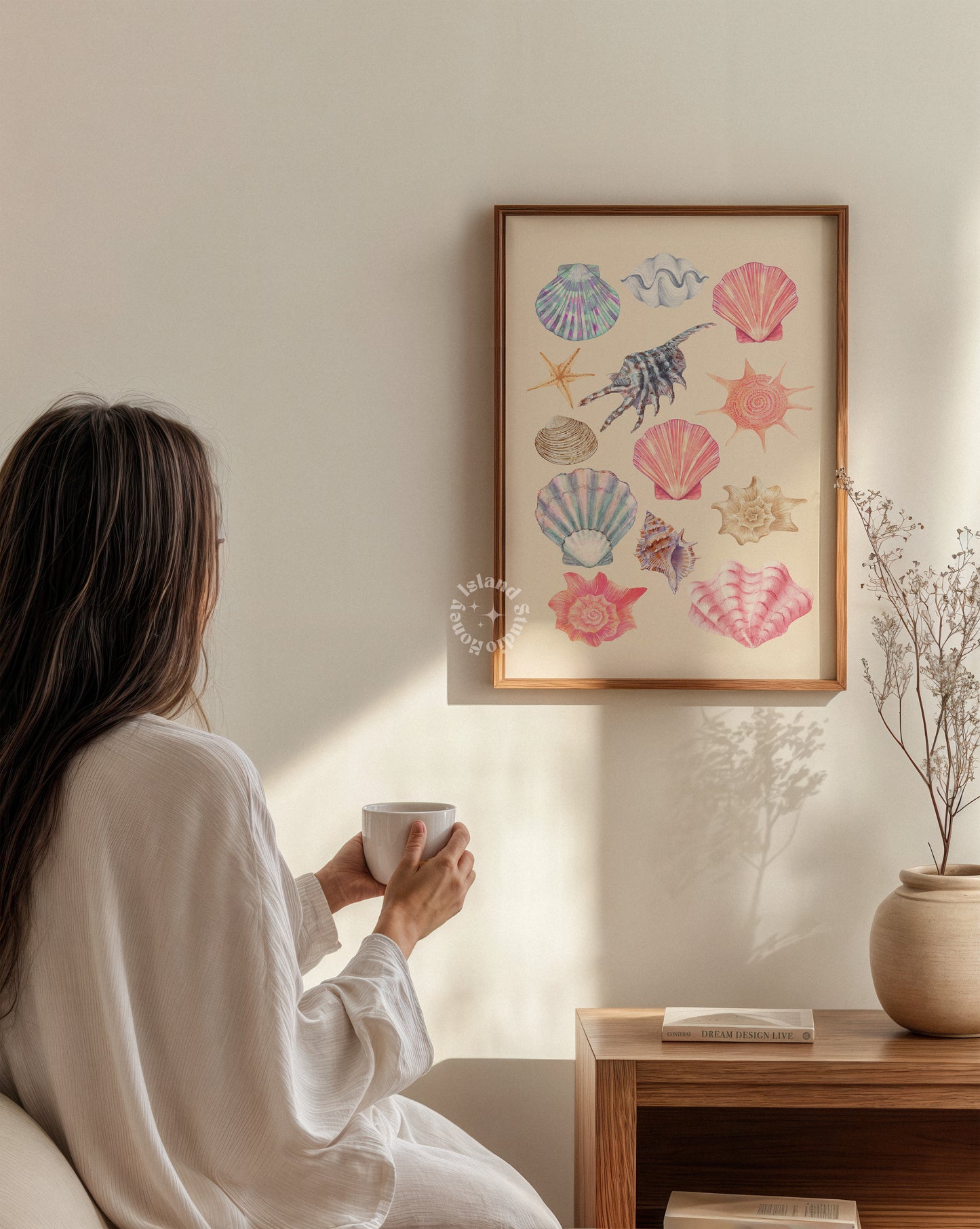 Woman in a white robe holding a mug, sitting in a room with a framed artwork of colorful seashells on the wall.