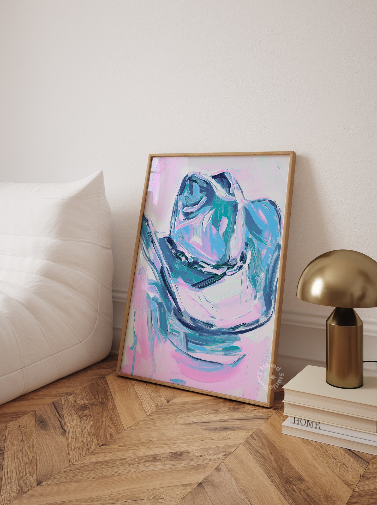 Abstract painting in a gold frame on a wall next to a lamp and books on a wooden floor.