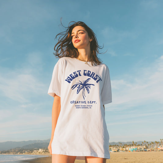 Woman wearing a 'West Coast Creative Dept.' t-shirt on a beach with a clear sky.