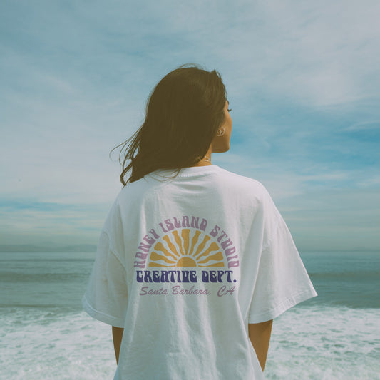 Person wearing a white t-shirt with text and graphics on the back, standing on a beach.