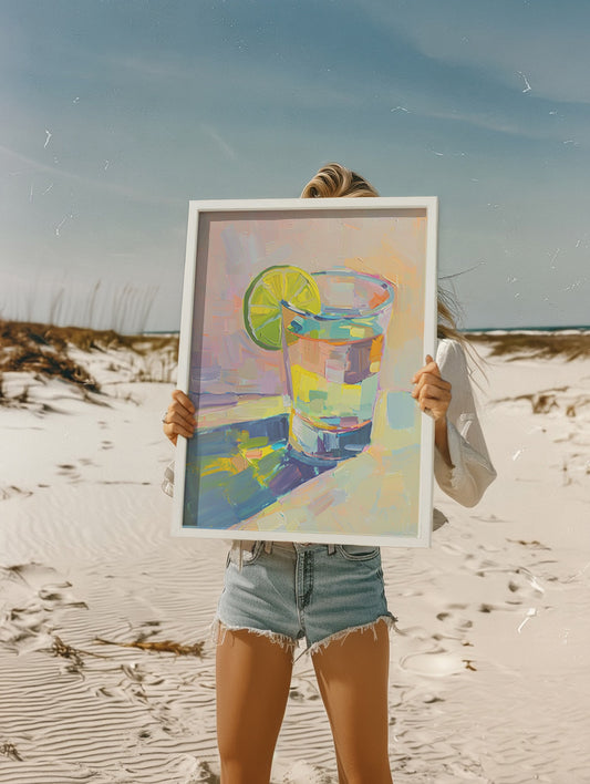 Person holding a framed painting of a colorful tequila drink with a lime on a beach.