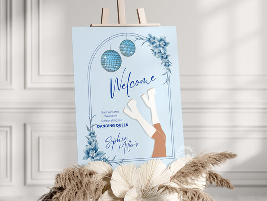 Light blue Mamma Mia-themed welcome sign for a bachelorette weekend party, elegantly displayed on an easel in a sunlit room with a white wall background. The sign features a Mediterranean-inspired design, perfect for a fun and stylish celebration.