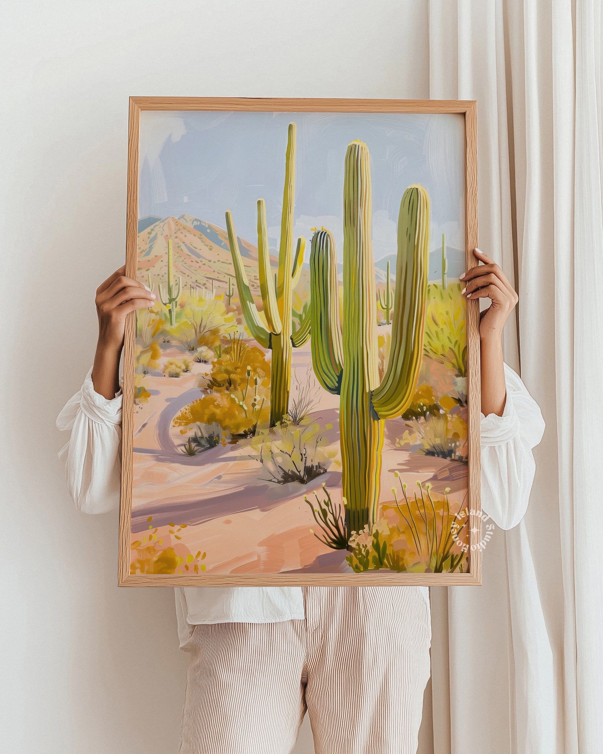 Person holding a framed painting of a desert landscape with cacti.