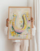 Person holding a framed artwork of a colorful horseshoe with flowers against a white wall.