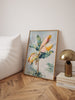 Framed floral artwork on a wall next to a gold lamp and books on a wooden floor.