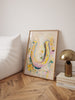 Framed artwork of a colorful horseshoe on a wall next to a lamp and books.