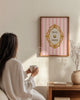 Woman in a white robe holding a cup, sitting in front of a decorative wall art piece.