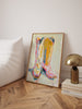 Framed western art piece on a wall next to a lamp and books on a wooden floor.
