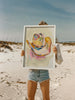 Person holding a framed abstract painting on a beach