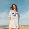Woman wearing a 'West Coast Creative Dept.' t-shirt on a beach with a clear sky.