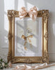 Decorative gold frame with a pink bow and floral arrangement on a white wall.