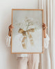 Person holding a framed artwork with a floral design and bow against a white wall.