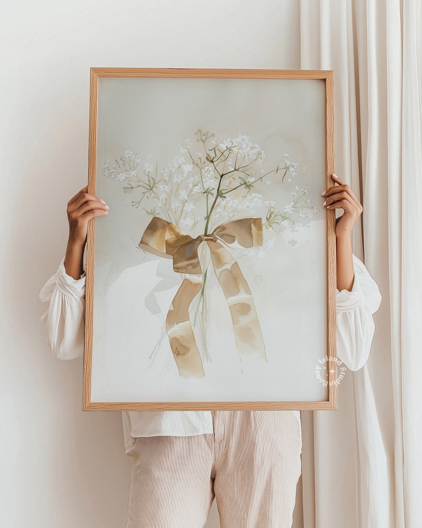 Person holding a framed artwork with a floral design and bow against a white wall.