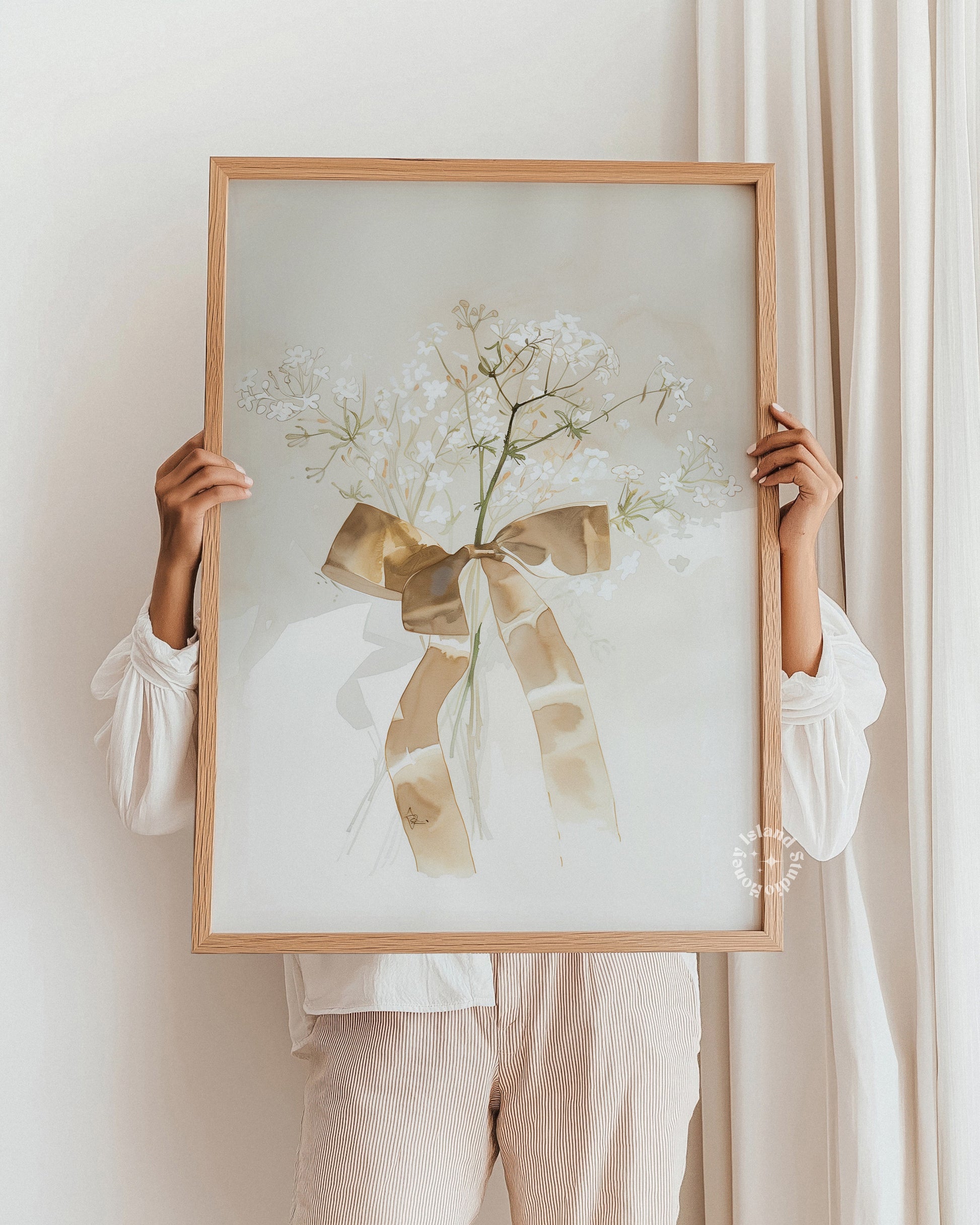 Person holding a framed artwork with a floral design and bow against a white wall.