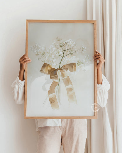 Person holding a framed artwork with a floral design and bow against a white wall.