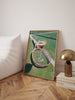 Framed artwork of a tennis racket and ball on a wall next to a lamp and books.