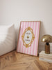 Framed pink and white striped poster with gold decorative elements on a wooden floor.