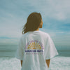 Person wearing a white t-shirt with text and graphics on the back, standing on a beach.