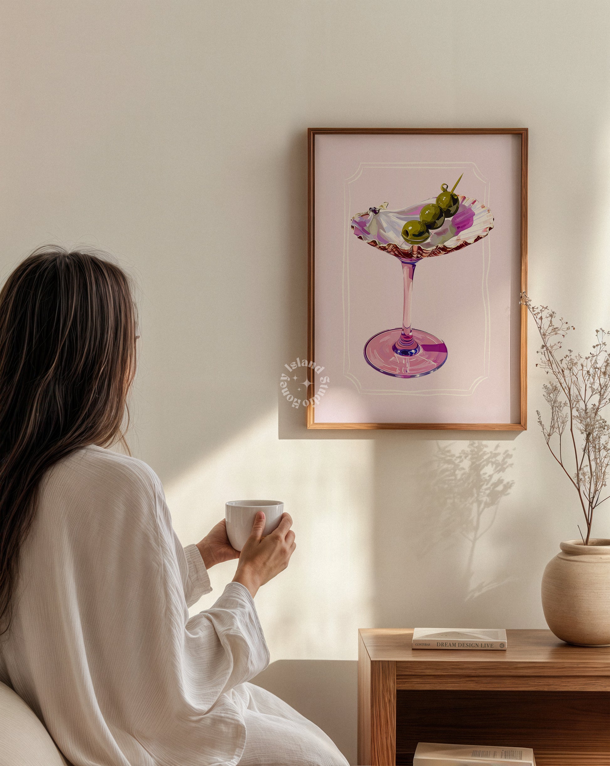Woman in a white robe holding a mug, sitting in a room with a framed artwork of a cocktail on the wall.