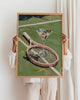 Person holding a framed painting of a tennis racket and martini glass against a white wall.