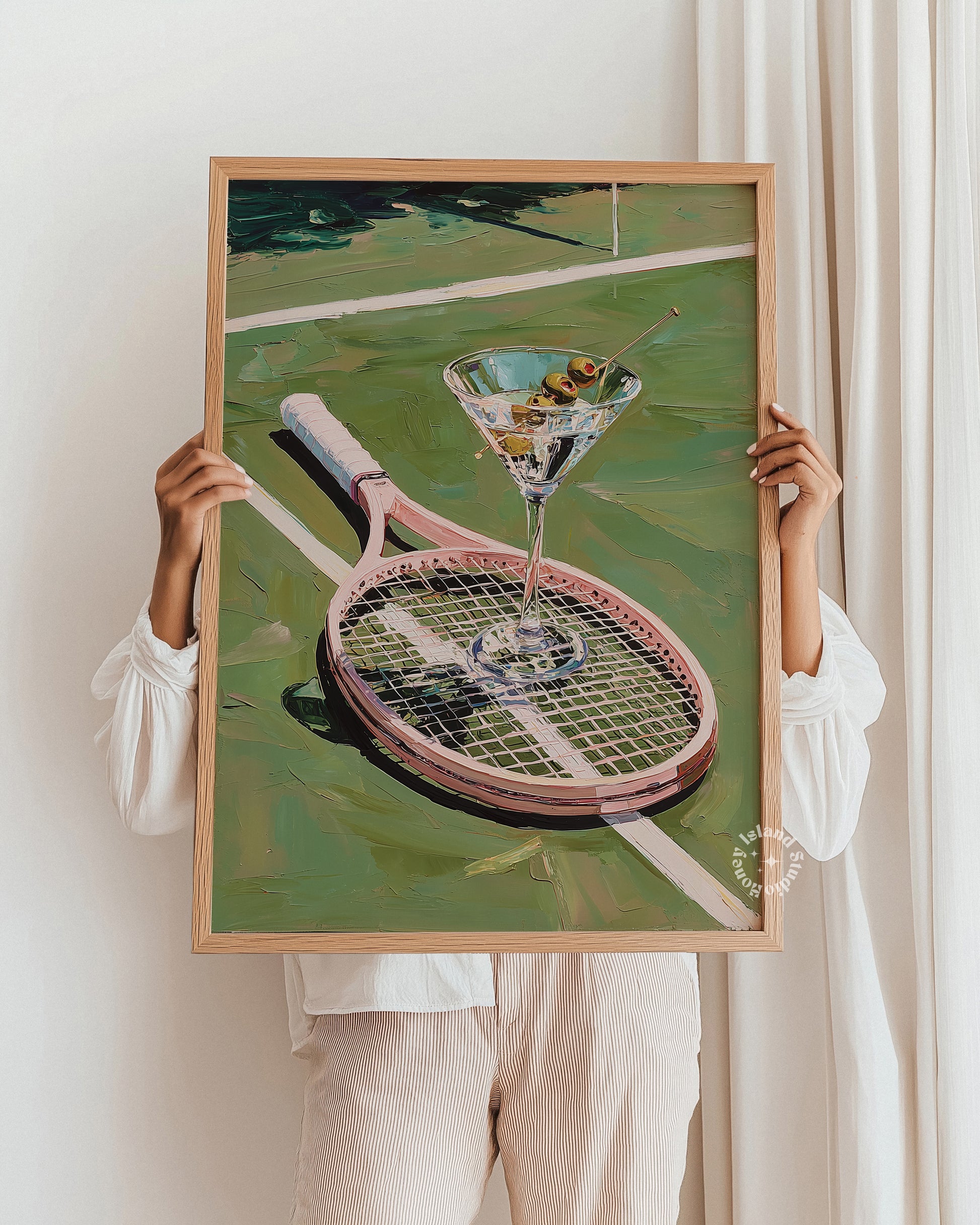 Person holding a framed painting of a tennis racket and martini glass against a white wall.