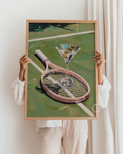 Person holding a framed painting of a tennis racket and martini glass against a white wall.