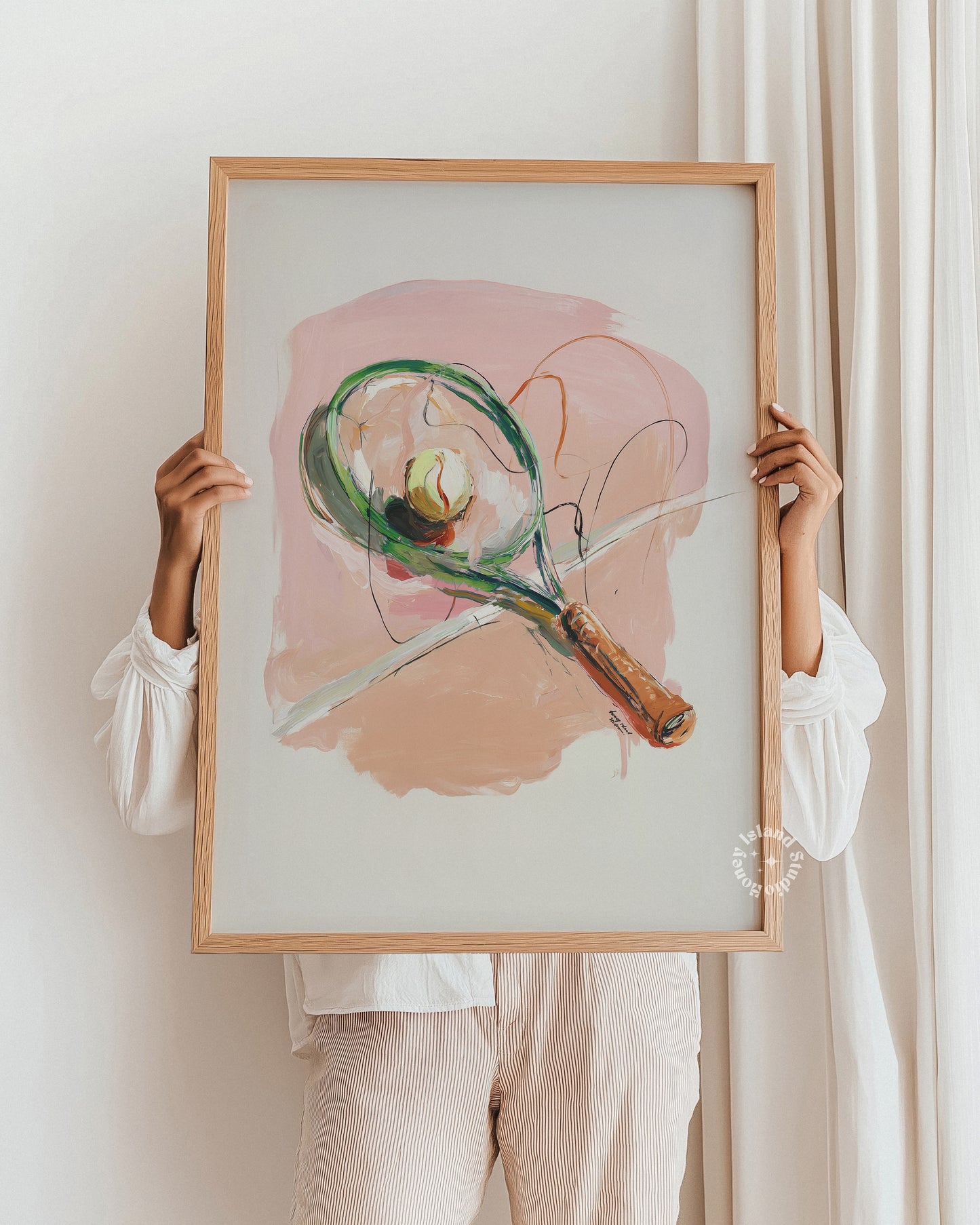 Person holding a framed abstract painting of a tennis racket against a white wall.