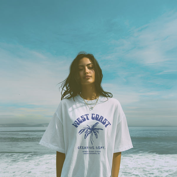 Woman wearing a 'West Coast' t-shirt with a palm tree design on a beach.